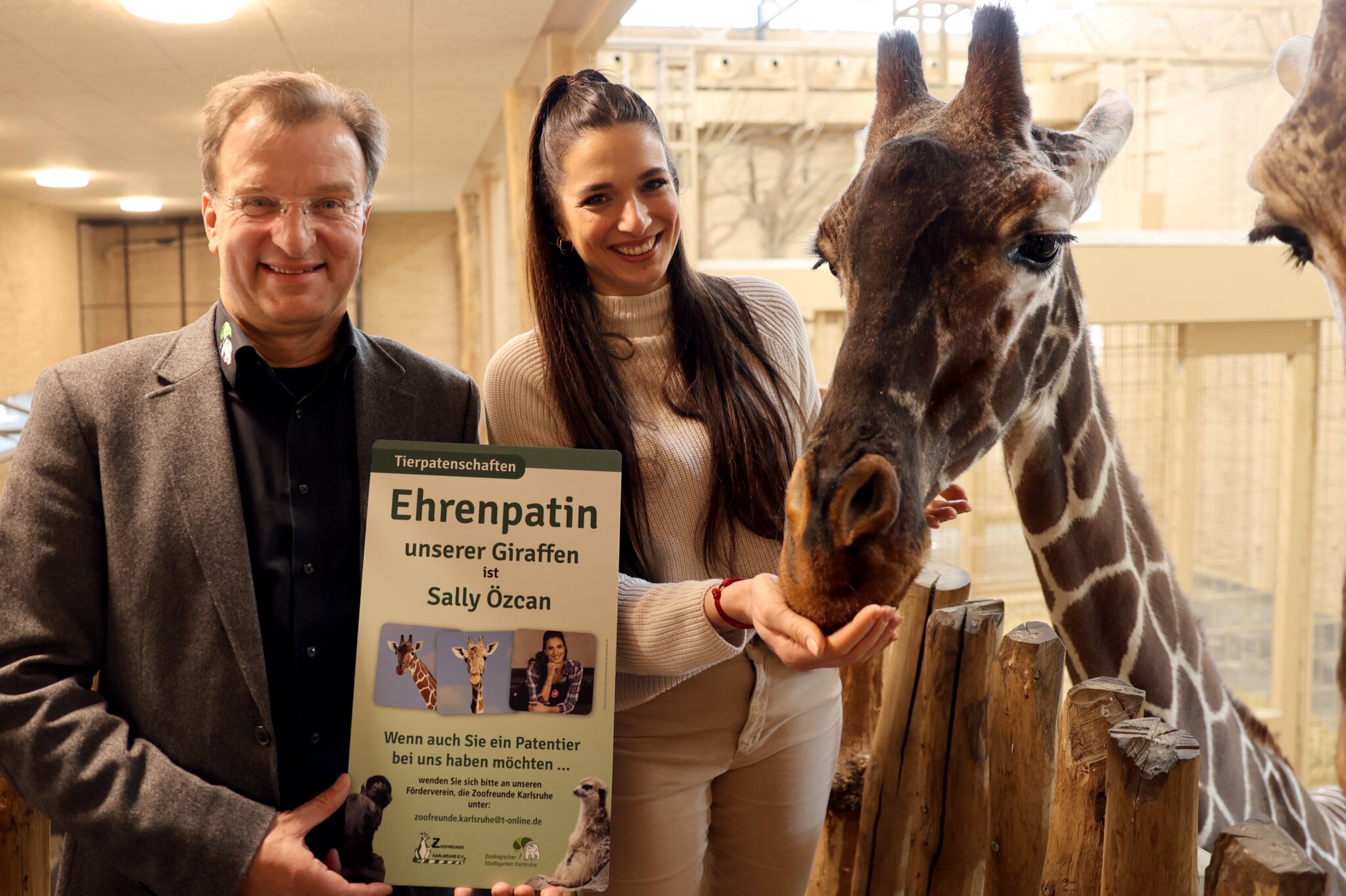 Ein Mann und eine Frau stehen in einem Zoogehege neben einer Giraffe und halten ein Sponsorenschild hoch.